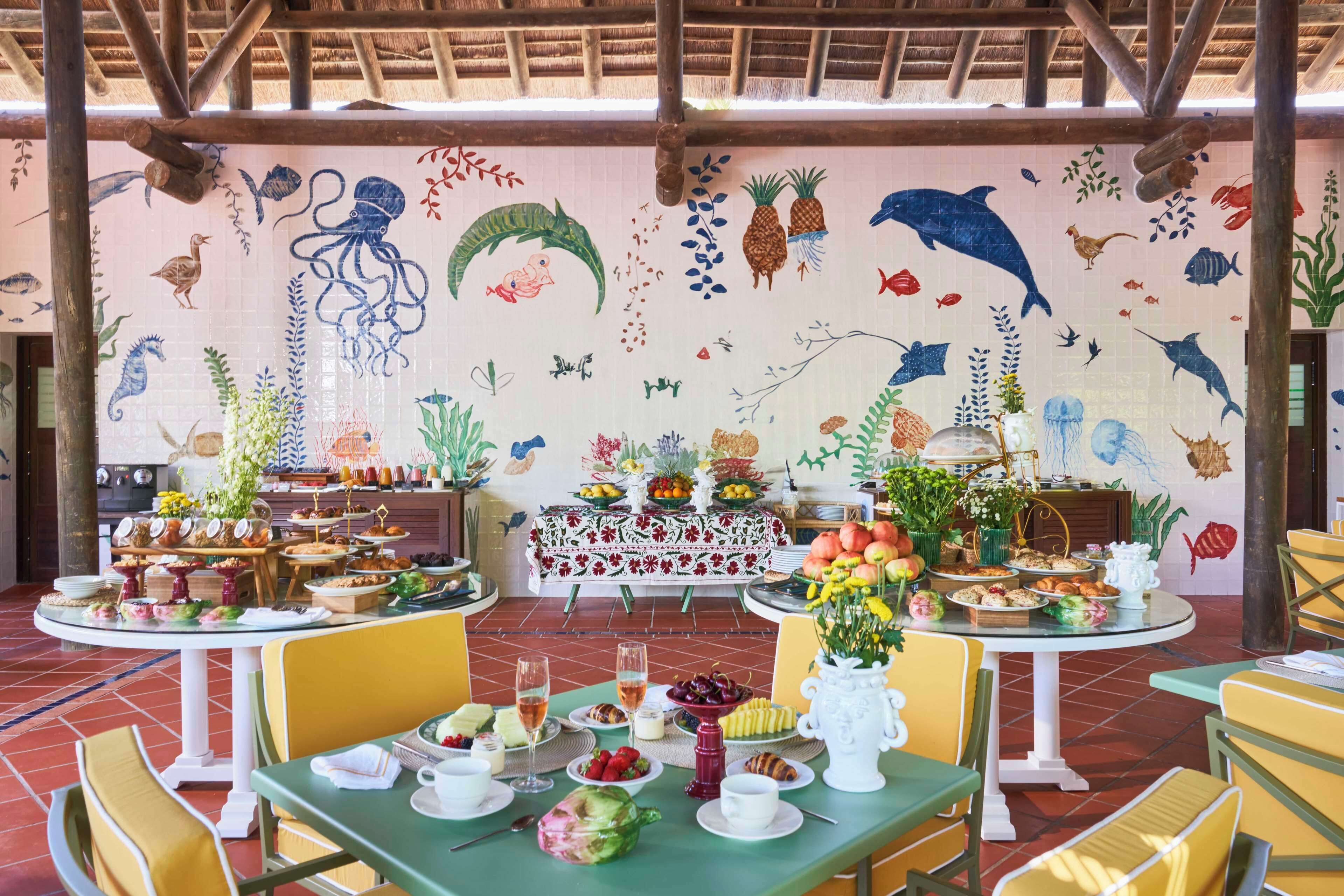 Un restaurante con un mural de vida marina en la pared, mesas dispuestas con comida y bebidas, y un techo tropical.