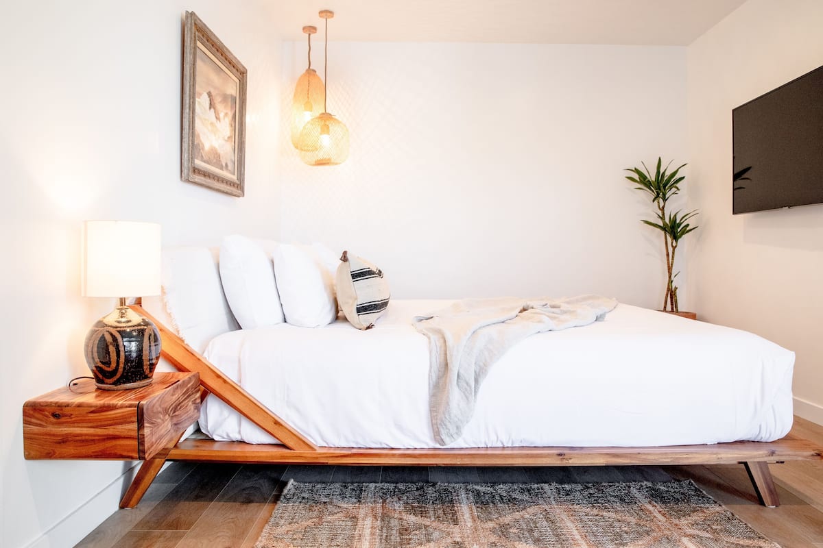 A bed with white bedding, a wooden nightstand with a lamp, a wall-mounted TV, and a potted plant.