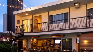 Zona junto a la piscina con bar y asientos exteriores, rodeada por un edificio con balcón y luces colgantes.
