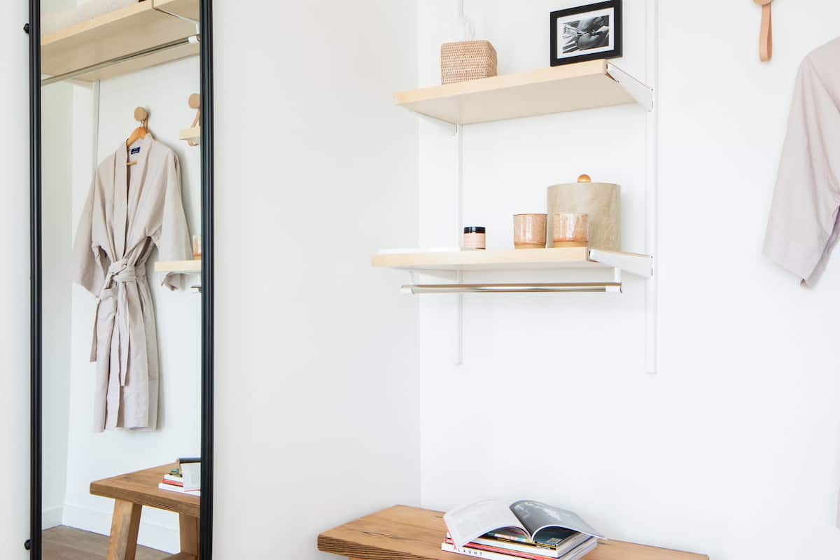 A bathroom with a wooden bench, shelves with toiletries, a hanging robe, and a large mirror.