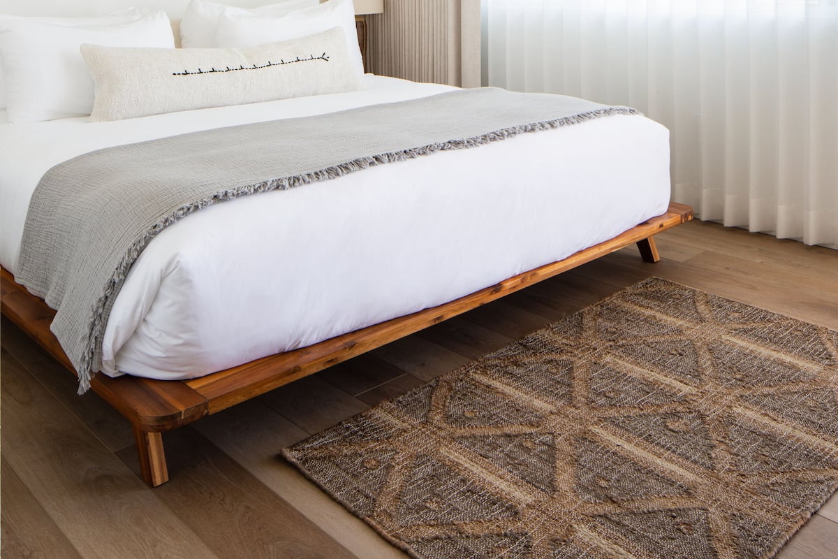 A modern bedroom with a wooden bed frame, white bedding, a patterned rug, a wall-mounted artwork, and sheer curtains.