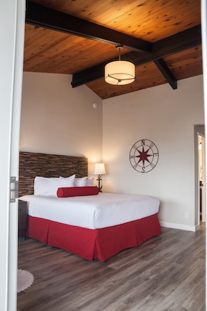 A bedroom with a wooden ceiling, a bed with white and red bedding, a wall-mounted compass, and a wooden floor. - The Landing at Morro Bay (Morro Bay)