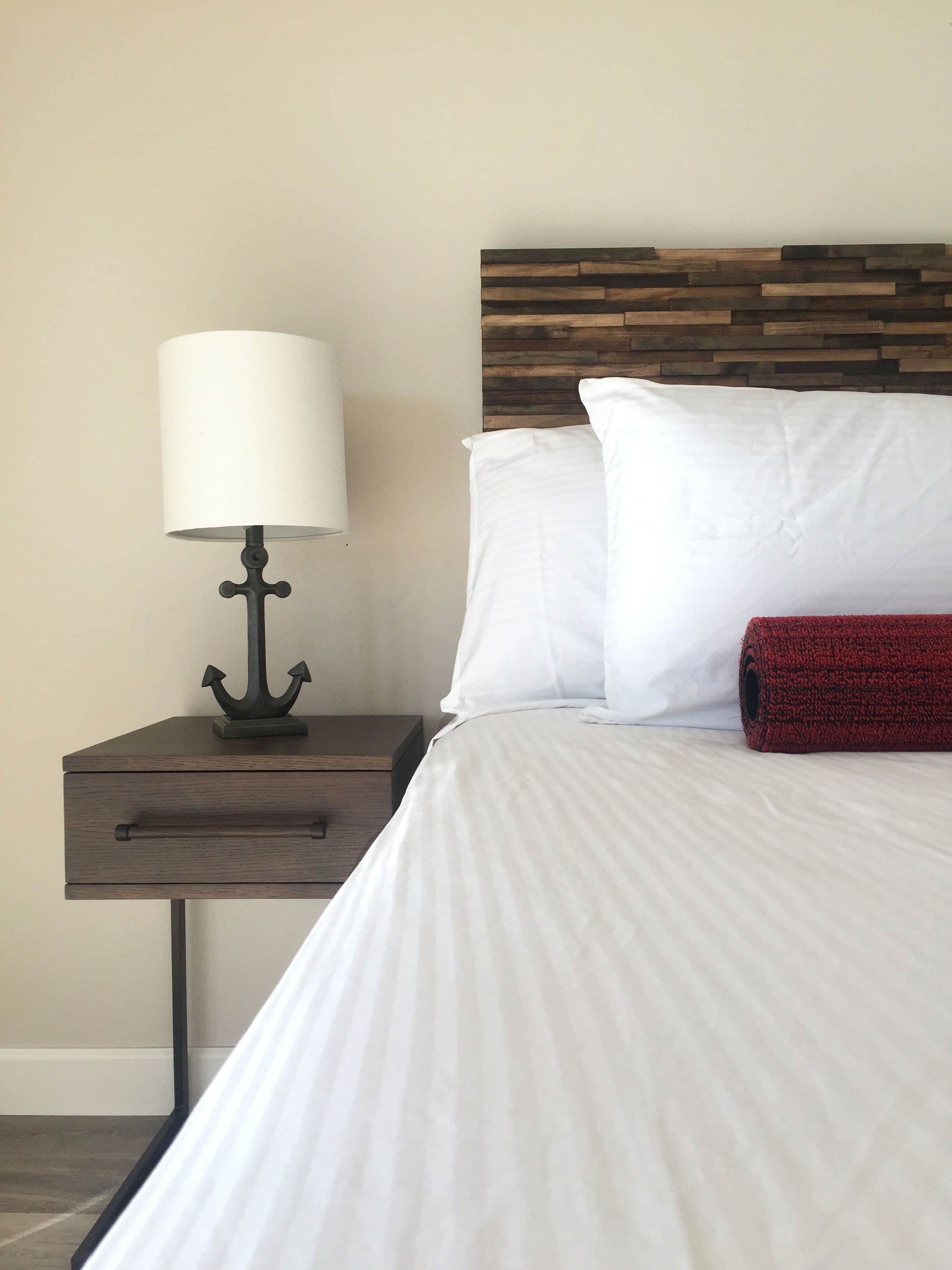 A neatly made bed with white linens and a red throw blanket, a wooden headboard, and a nightstand with a lamp and an anchor-shaped base.