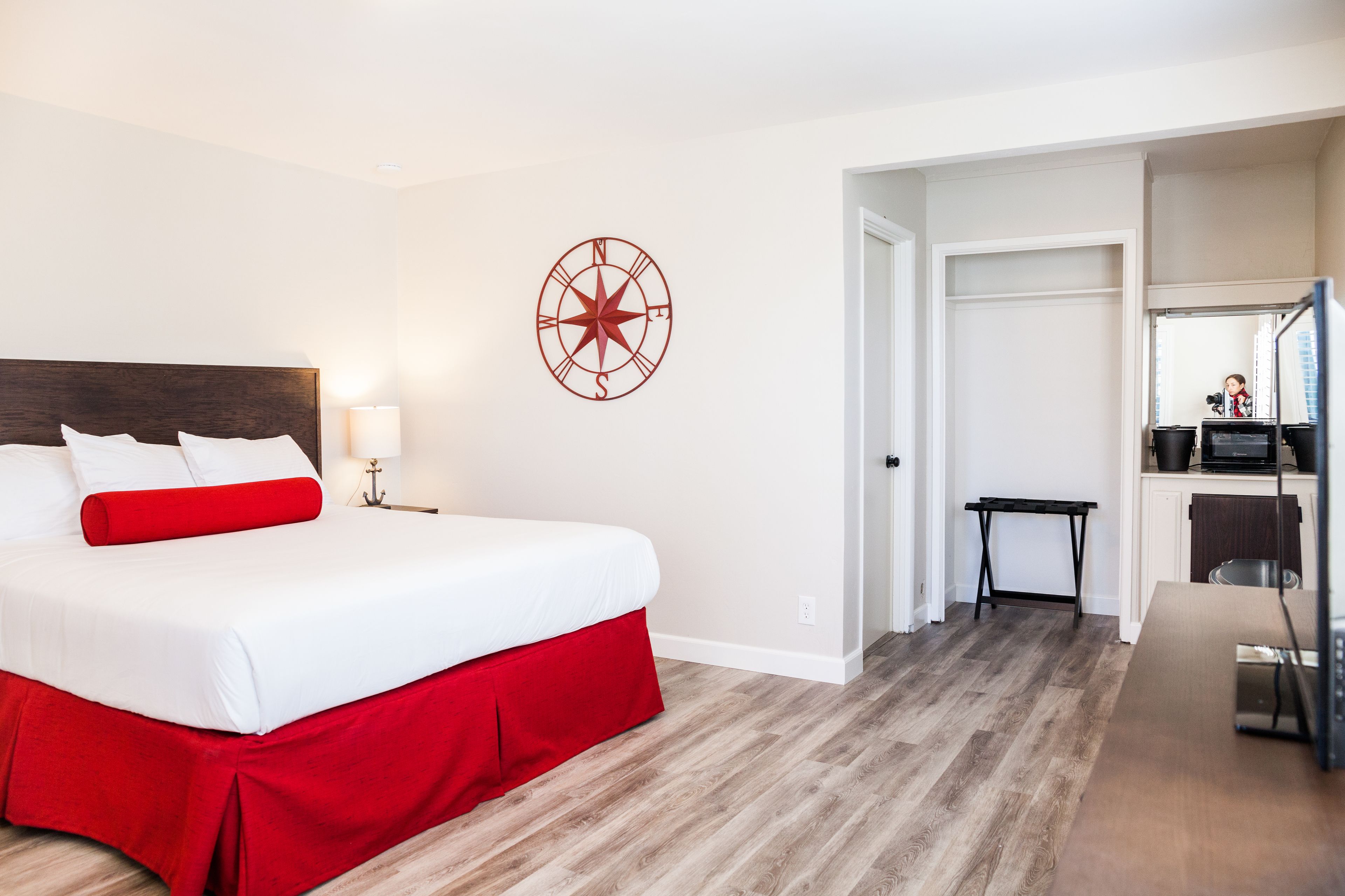 Une chambre d’hôtel avec un grand lit, un coussin rouge décoratif, une horloge murale, une petite table et un coin cuisine.
