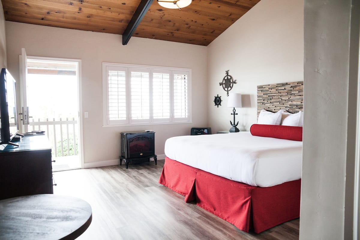 A bedroom with a large bed, a wooden ceiling, a window with blinds, a nightstand, and a desk with a lamp.