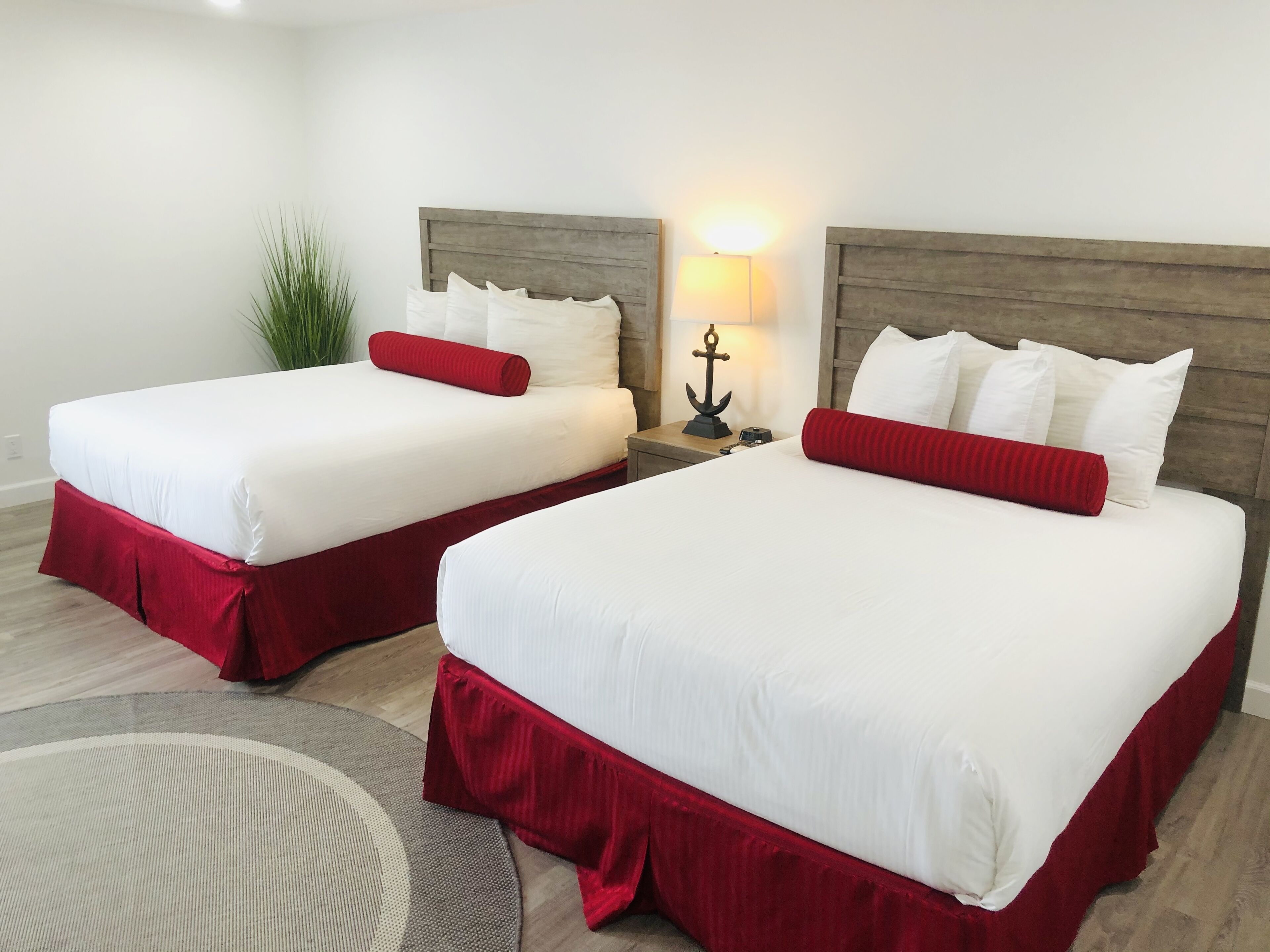 Two double beds with white linens and red accents, a nightstand with a lamp, and a potted plant.