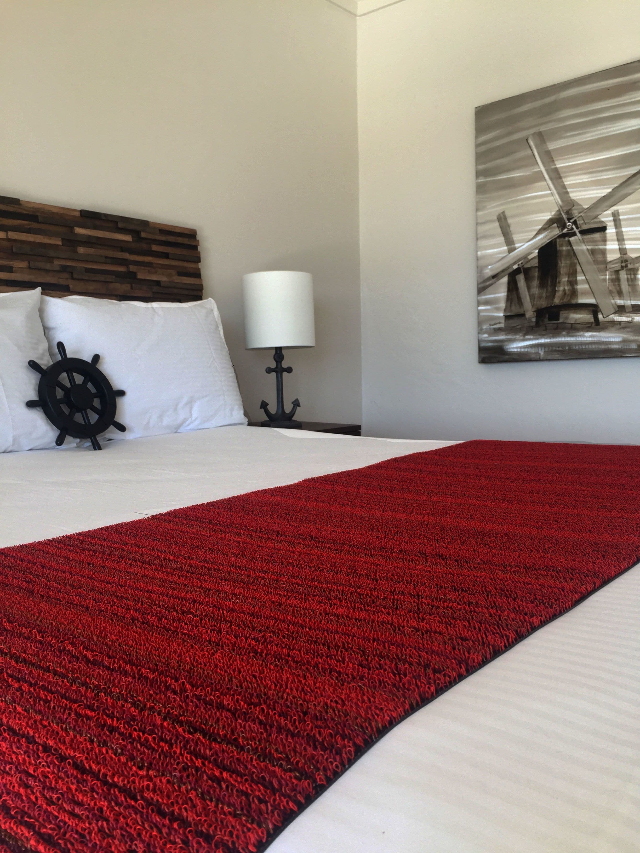A neatly made bed with a red blanket, a ship's wheel pillow, and a lamp on a bedside table.