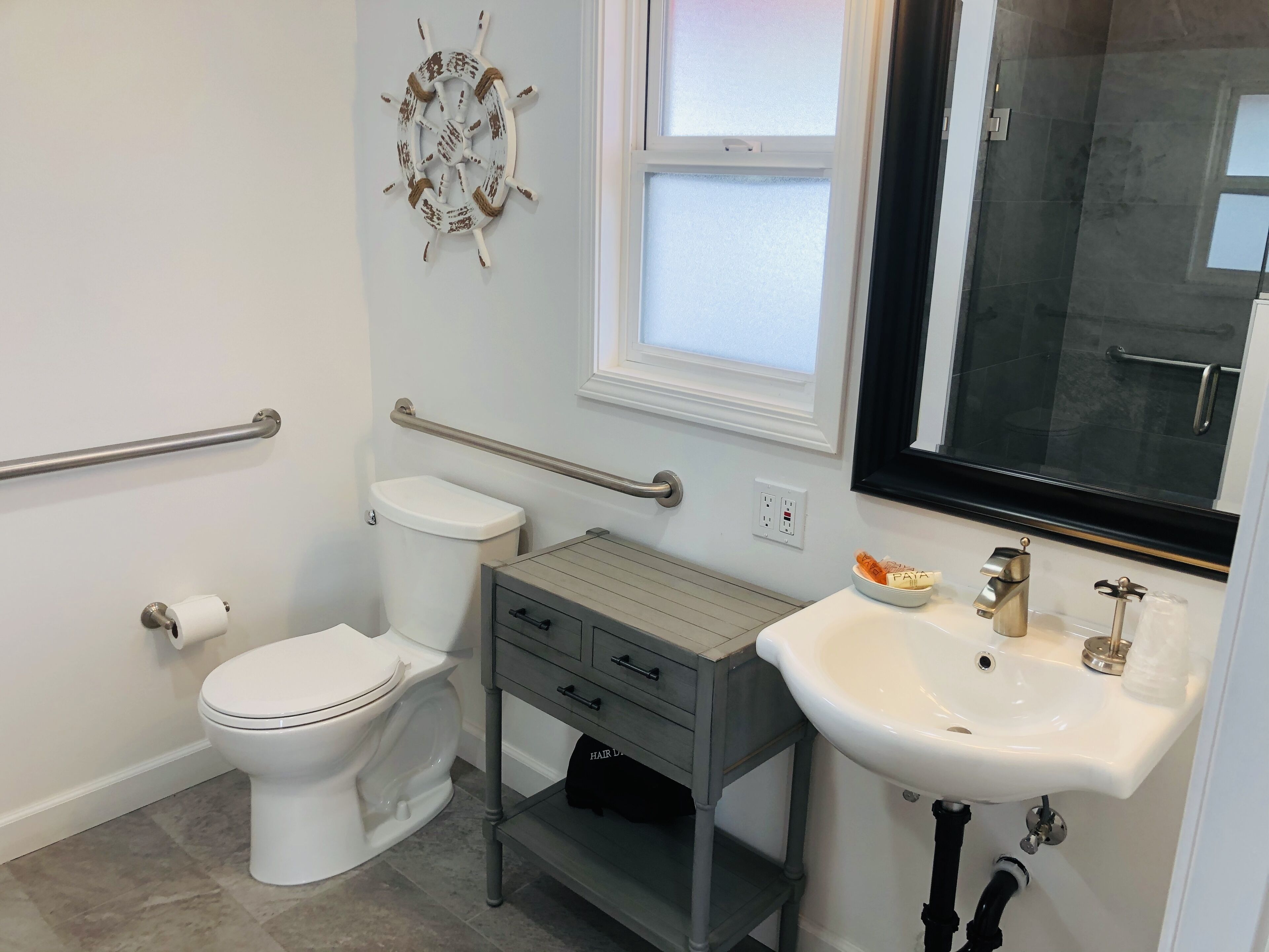 A bathroom with a toilet, a sink, a vanity, a window, and a ship's wheel decoration.