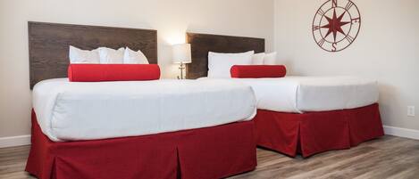 A hotel room with two beds, each with white bedding and red accents, a wooden headboard, a wall-mounted compass decoration, and a wooden floor.