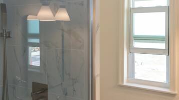 A glass-enclosed shower with a handheld showerhead, a small sink, and a window above the toilet.