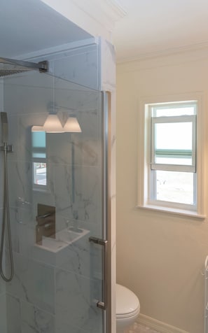 A glass-enclosed shower with a handheld showerhead, a small sink, and a window above the toilet.