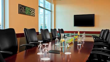 A conference room with a long table, black leather chairs, a flat-screen TV, and a window with blinds.