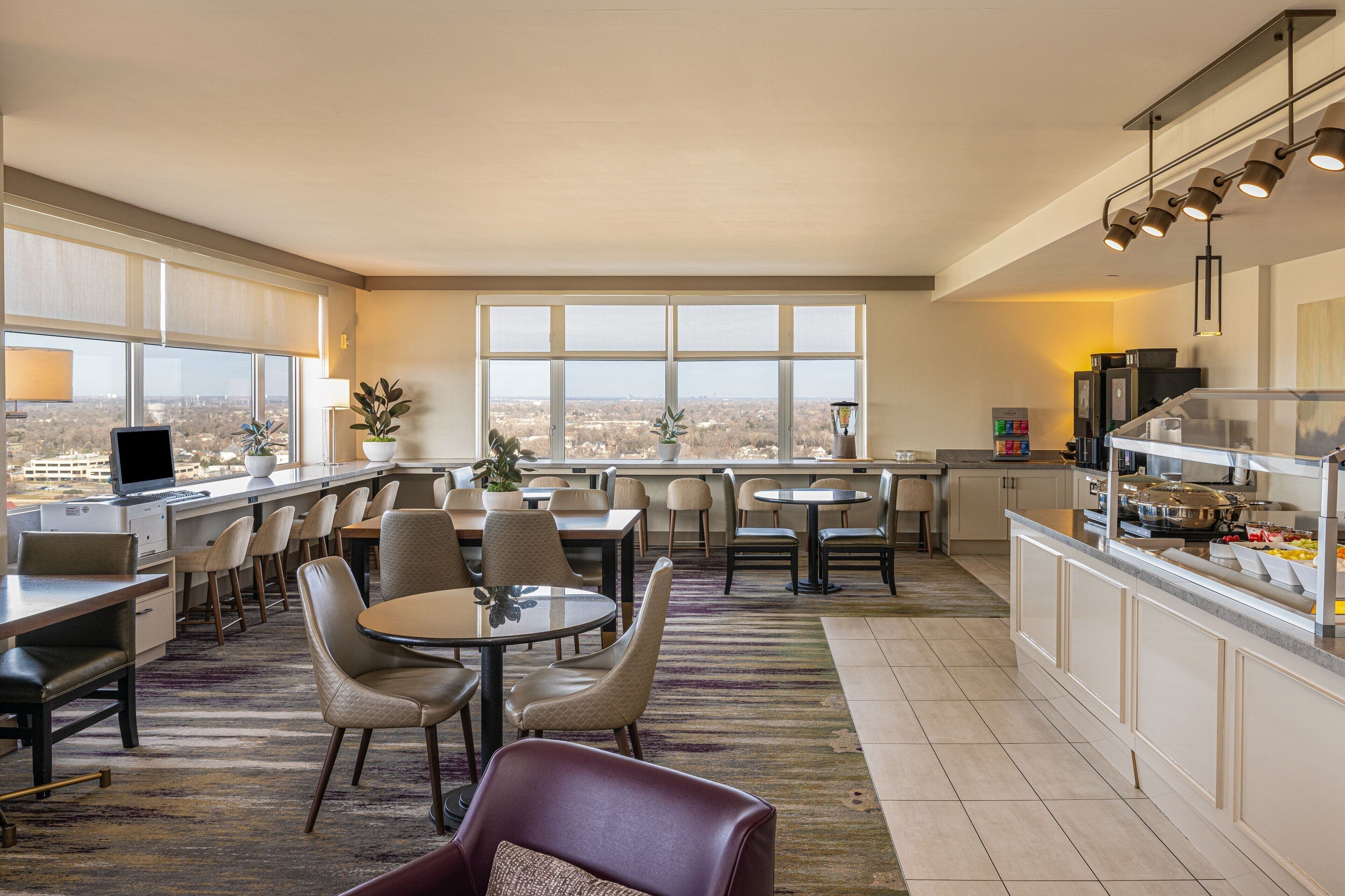 A modern hotel dining area with a panoramic city view, multiple seating options, and a buffet counter.