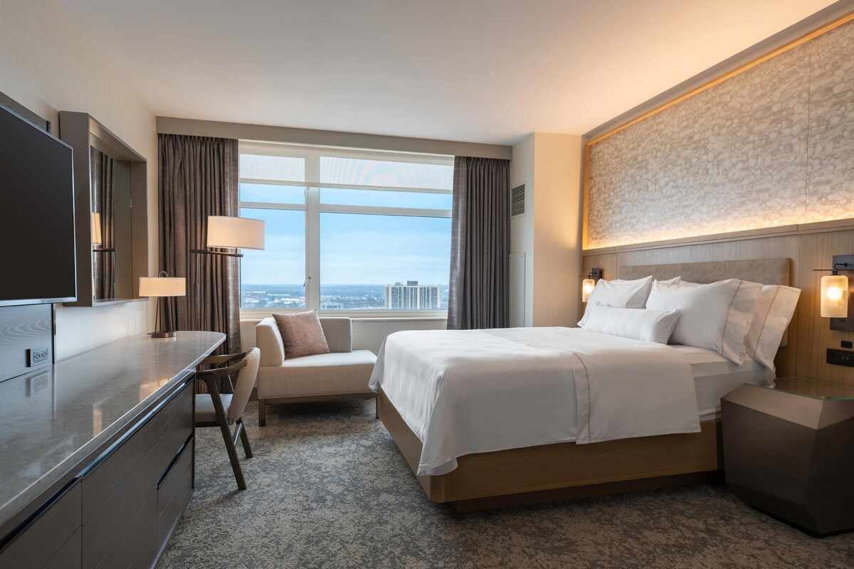 A hotel room with a bed, a desk, a chair, a TV, and a view of the city.
