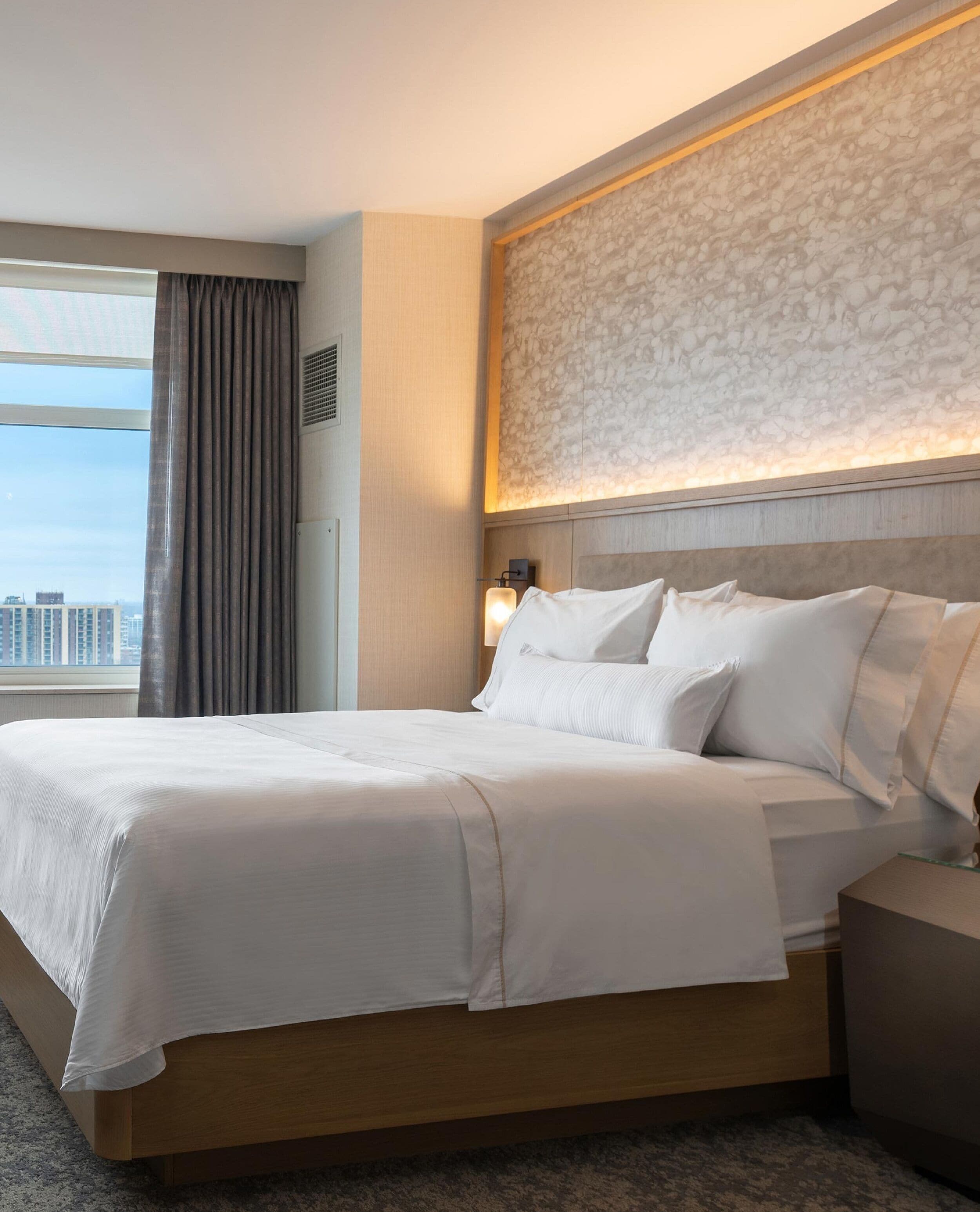 A hotel room with a large bed, a nightstand, a lamp, and a view of the city through a window with curtains.