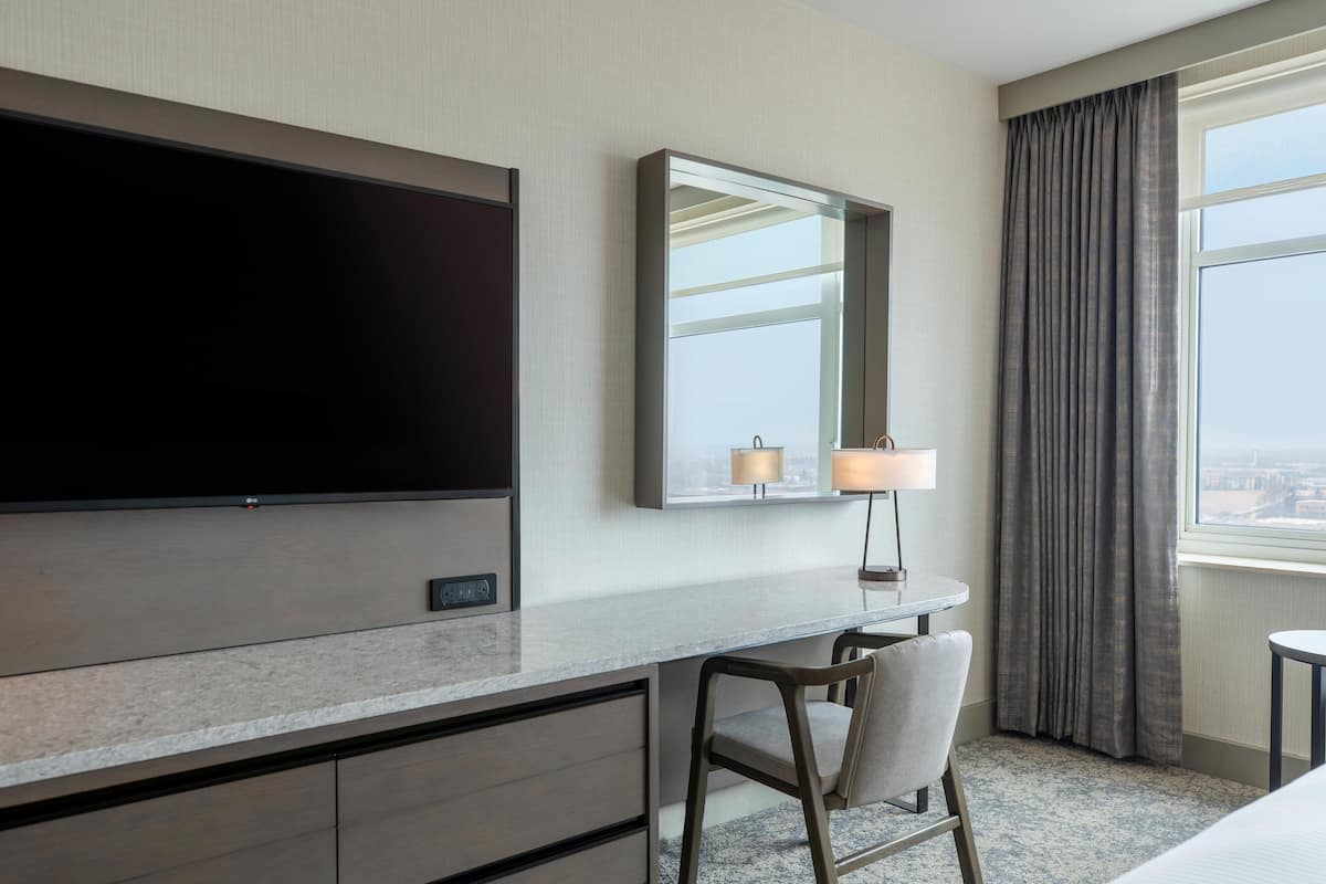 A hotel room with a large flat-screen TV, a desk with a lamp, a chair, and a view of the city through a window with curtains.