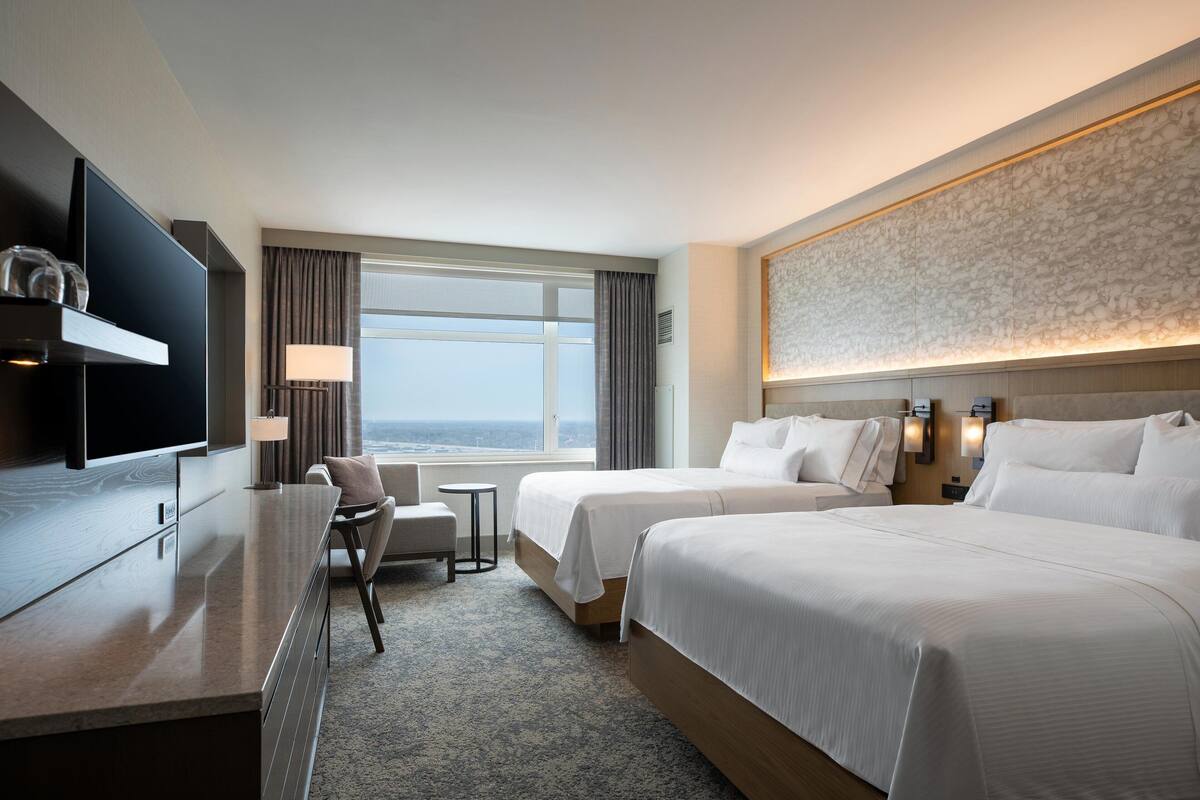A hotel room with two beds, a desk, a chair, a TV, and a large window with a view.