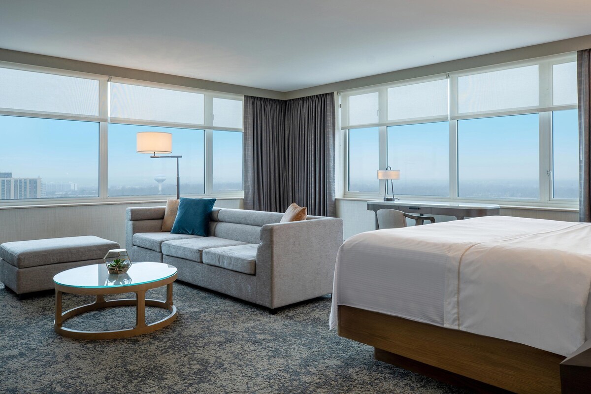 A hotel room with a bed, two sofas, a small round table, and large windows offering a city view.