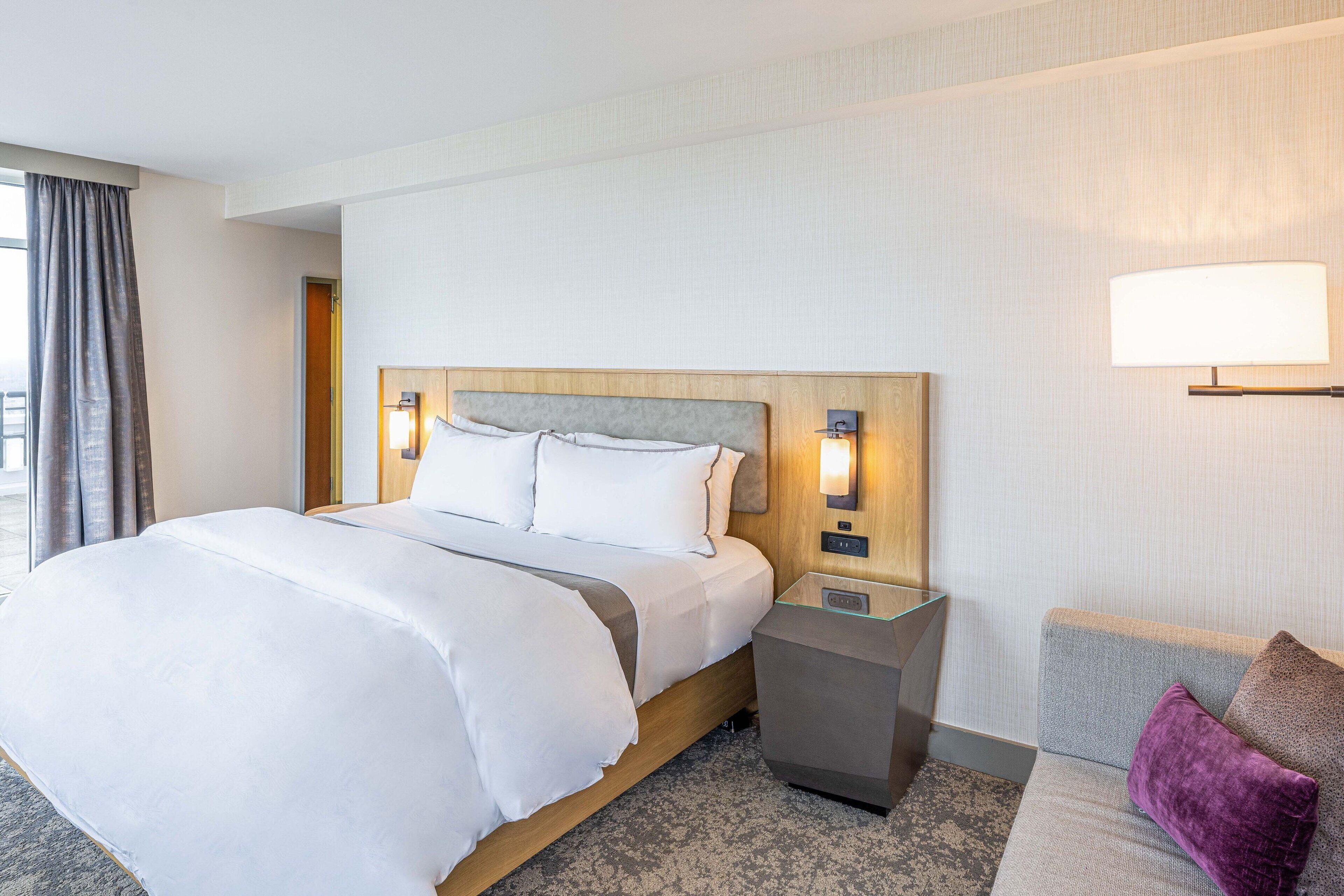 Habitación de hotel con una cama grande, mesitas de noche, un sofá y una ventana con cortinas.