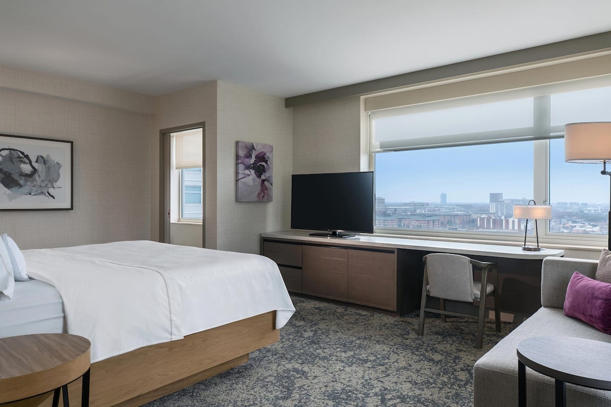 A hotel room with a bed, a desk, a TV, a chair, a lamp, and a view of the city.