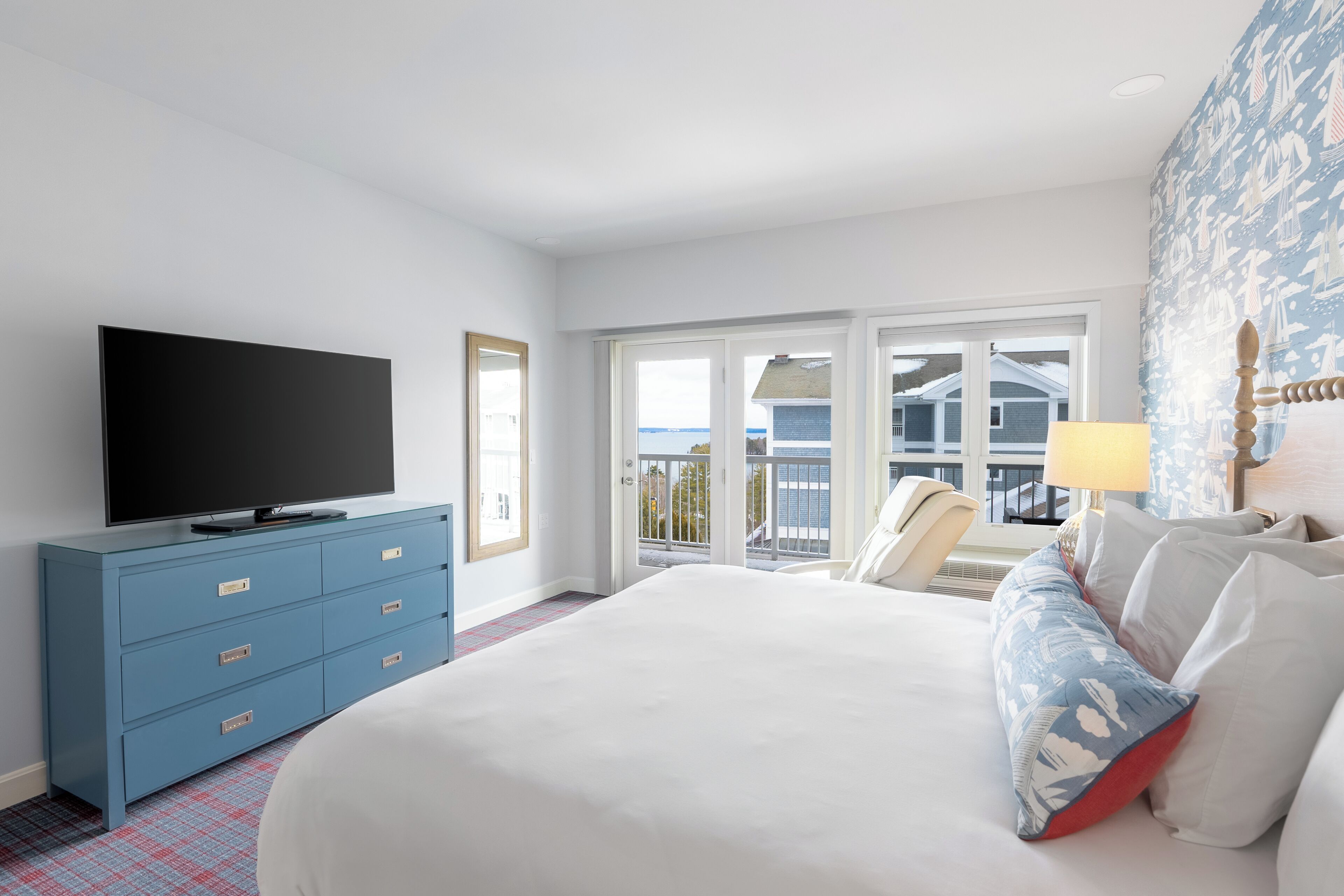 A hotel room with a large bed, a television on a blue dresser, a balcony with a view, and a patterned wall.