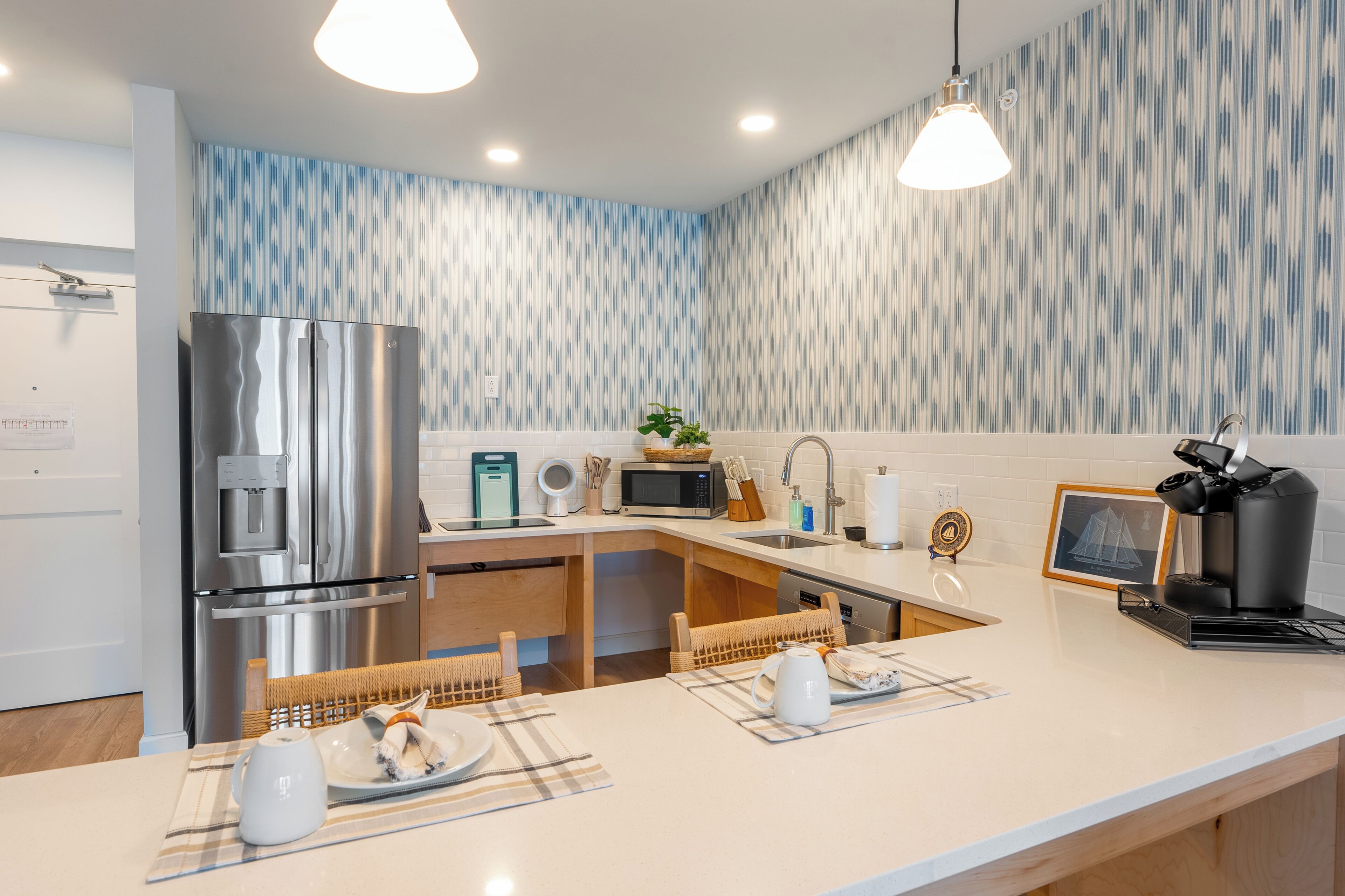 A modern kitchen with a stainless steel refrigerator, a microwave, and a coffee machine. There is a wooden dining table with a placemat and a white pitcher. The kitchen has a white countertop and a patterned backsplash.