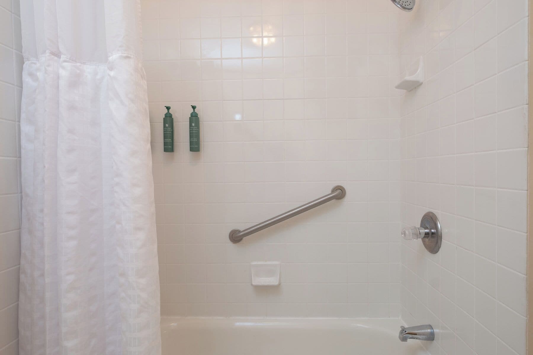 A shower with a grab bar, a handheld showerhead, and two bottles of shampoo on the wall.