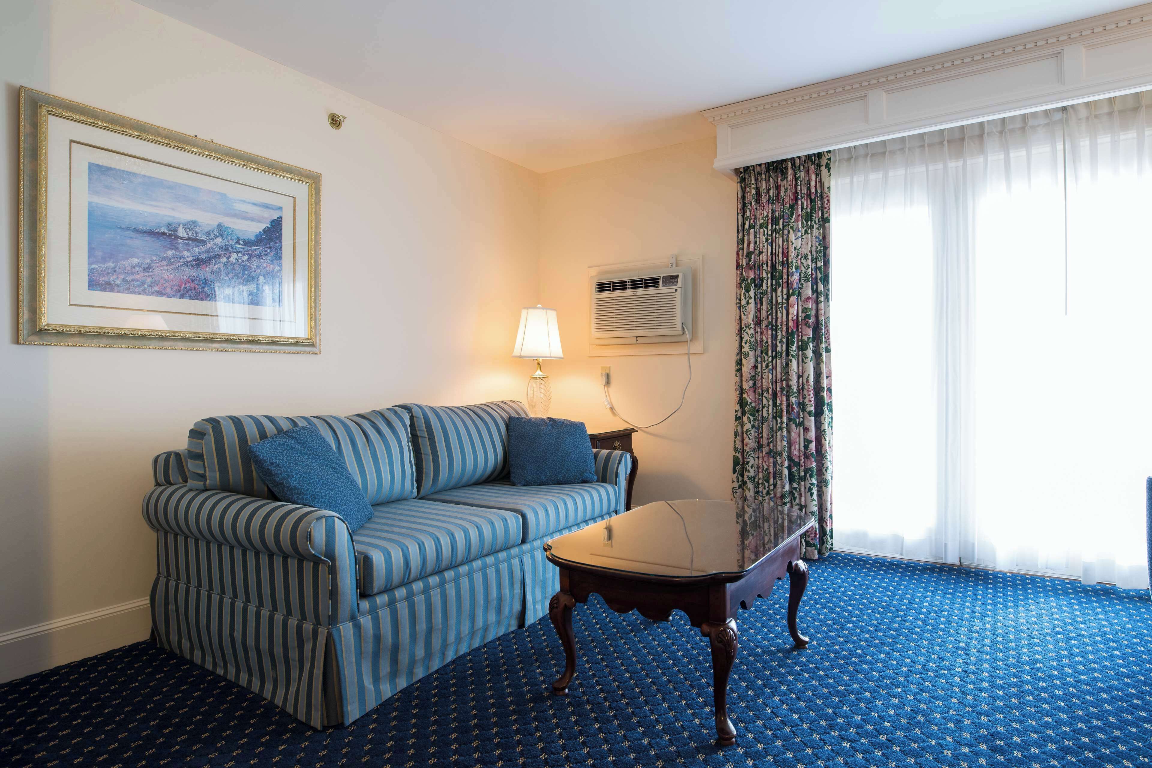 Une chambre d’hôtel avec un canapé à rayures, une petite table, une lampe, un appareil de climatisation et une grande fenêtre avec des rideaux.