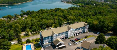 An aerial view of a hotel with a swimming pool, parking lot, and a view of the surrounding water and greenery.
