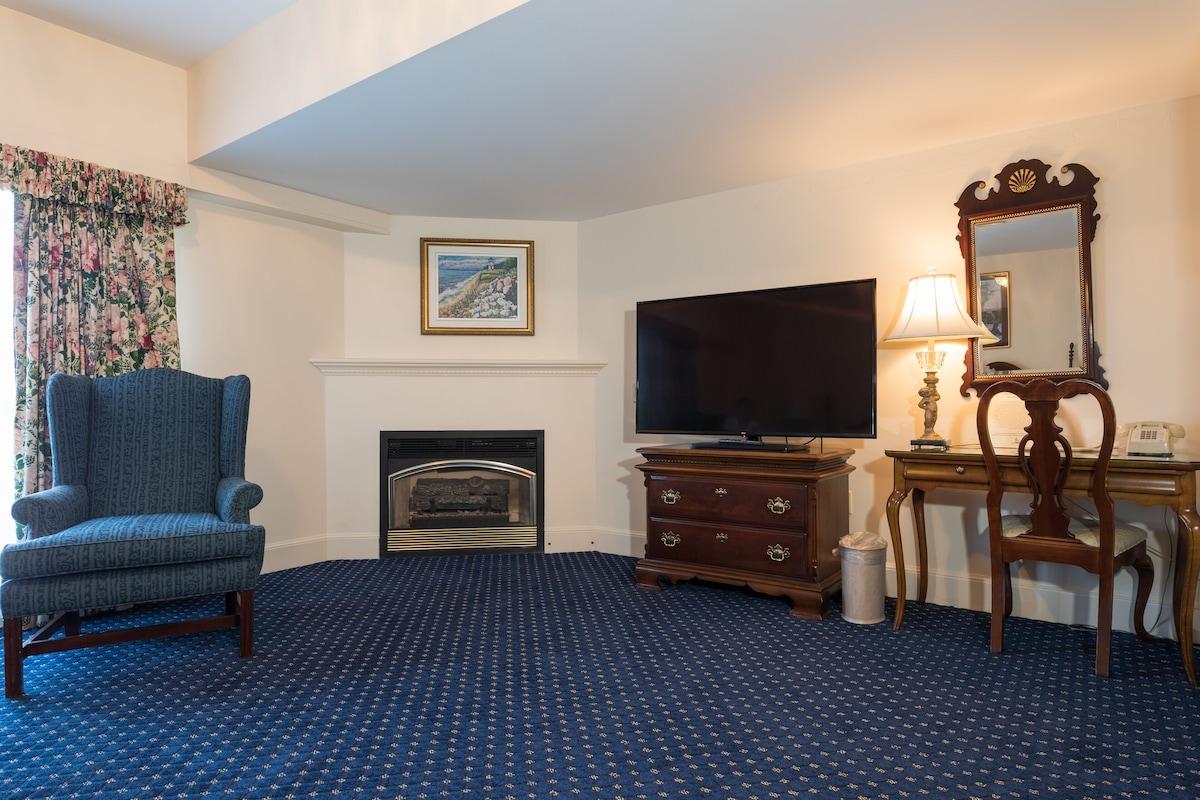A hotel room with a blue carpet, a fireplace, a television, a wooden dresser, and a chair.