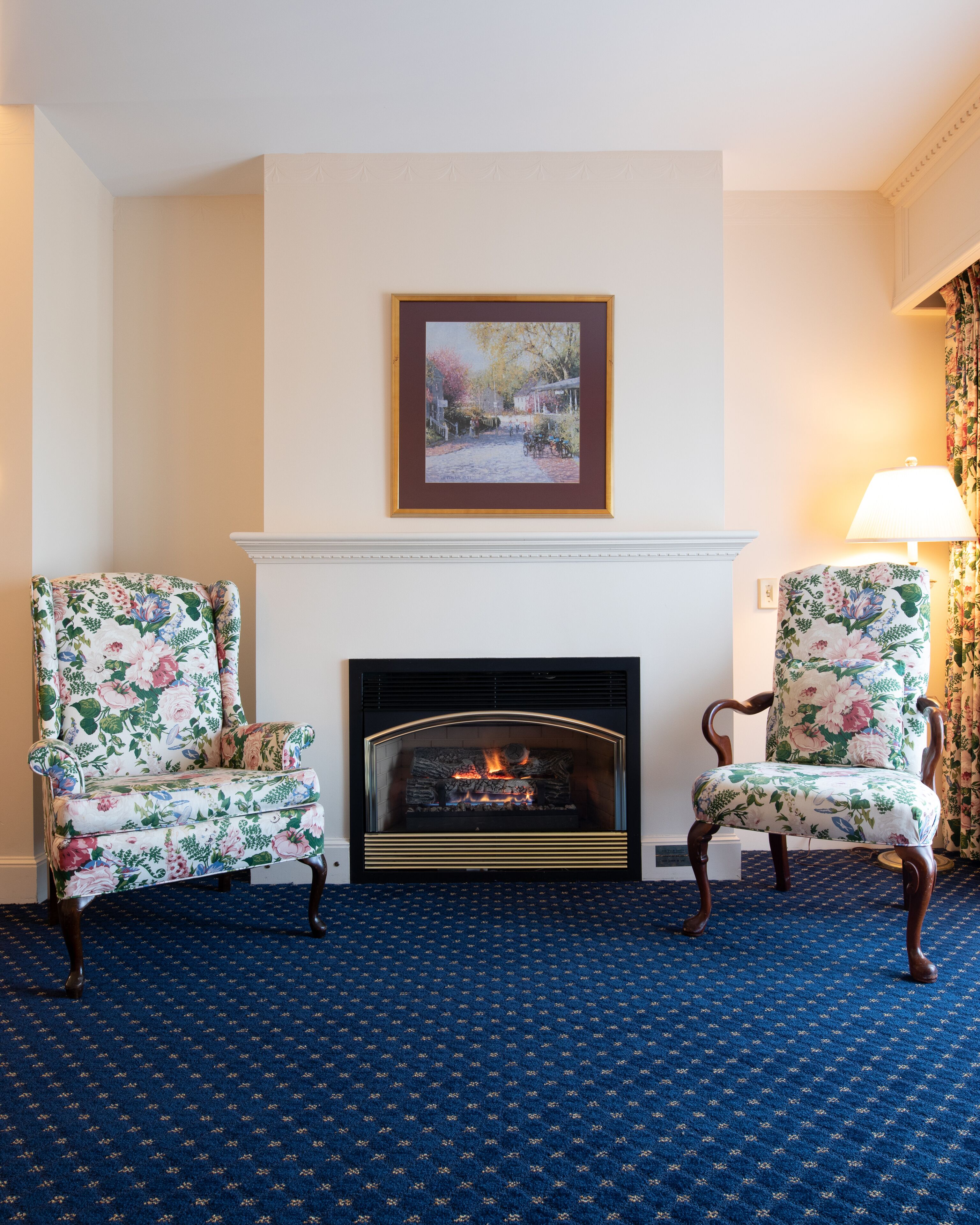 A room with a fireplace, two floral armchairs, a framed picture above the fireplace, and a patterned carpet.