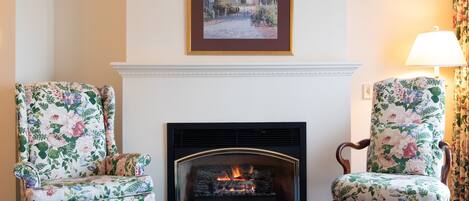A room with a fireplace, two floral armchairs, a framed picture above the fireplace, and a patterned carpet.