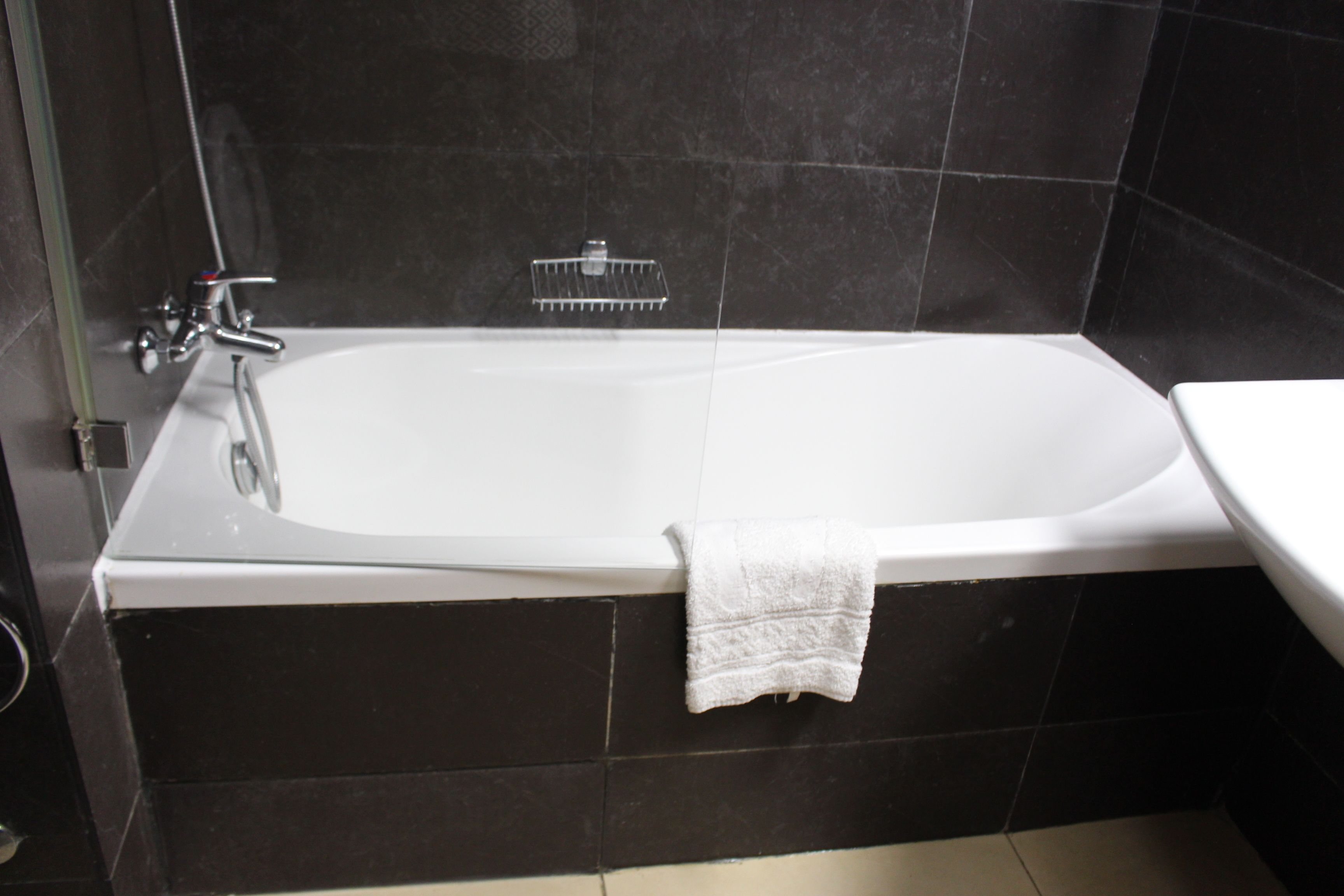 A modern bathroom with a white bathtub, a white sink, and dark tiled walls.