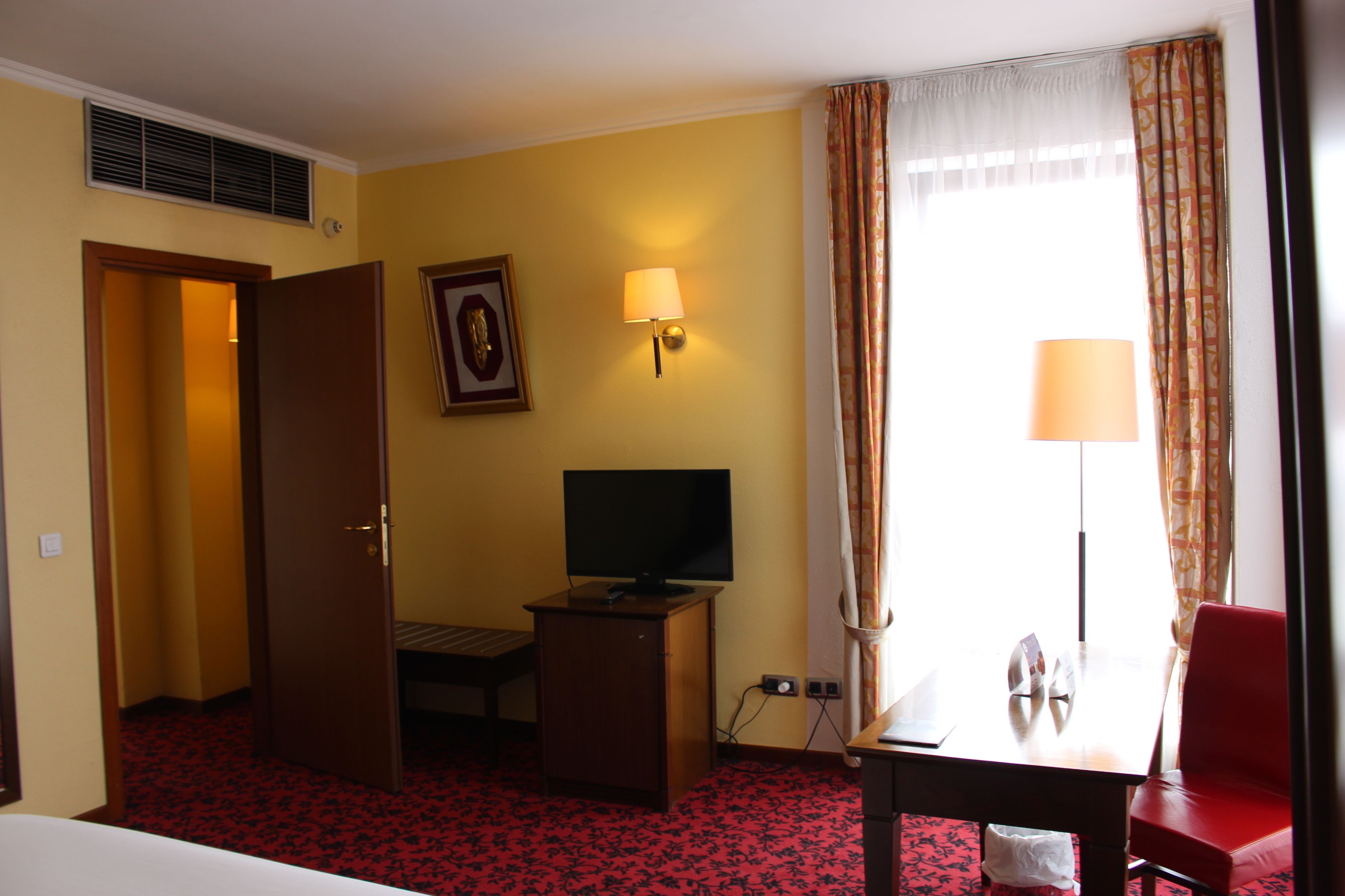 A hotel room with a bed, a desk with a television, a red chair, a lamp, and a window with curtains.