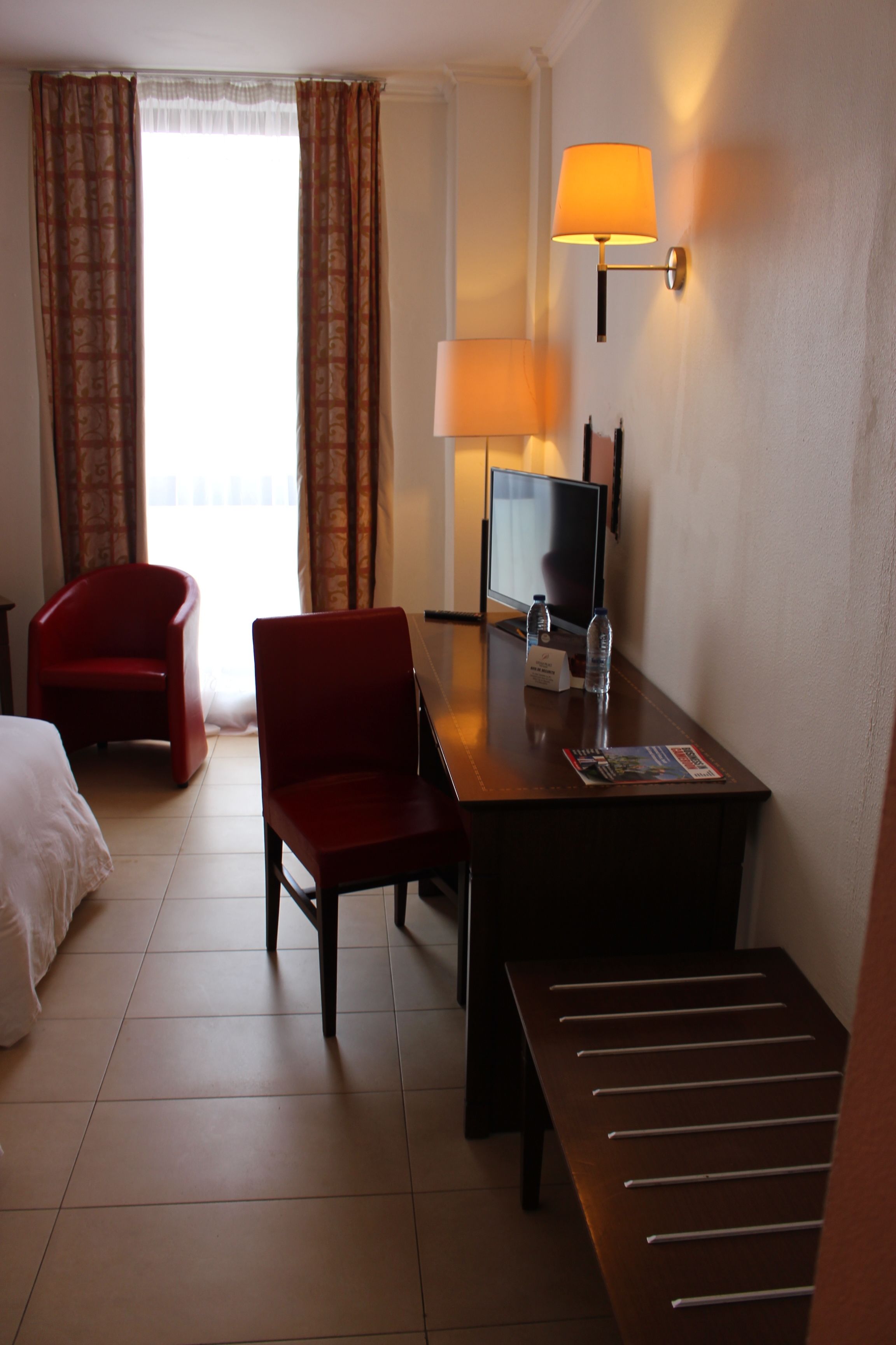 A hotel room with a bed, a desk with a lamp, a chair, a TV, and a window with curtains.