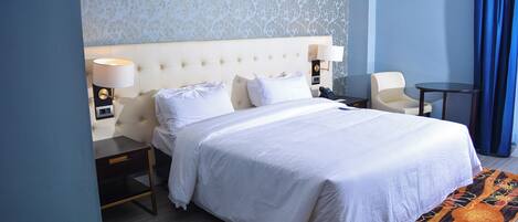 Una cama bien tendida con sábanas blancas, una mesita de noche con una lámpara y una silla en una habitación de hotel con papel tapiz estampado y una pared azul de acento.