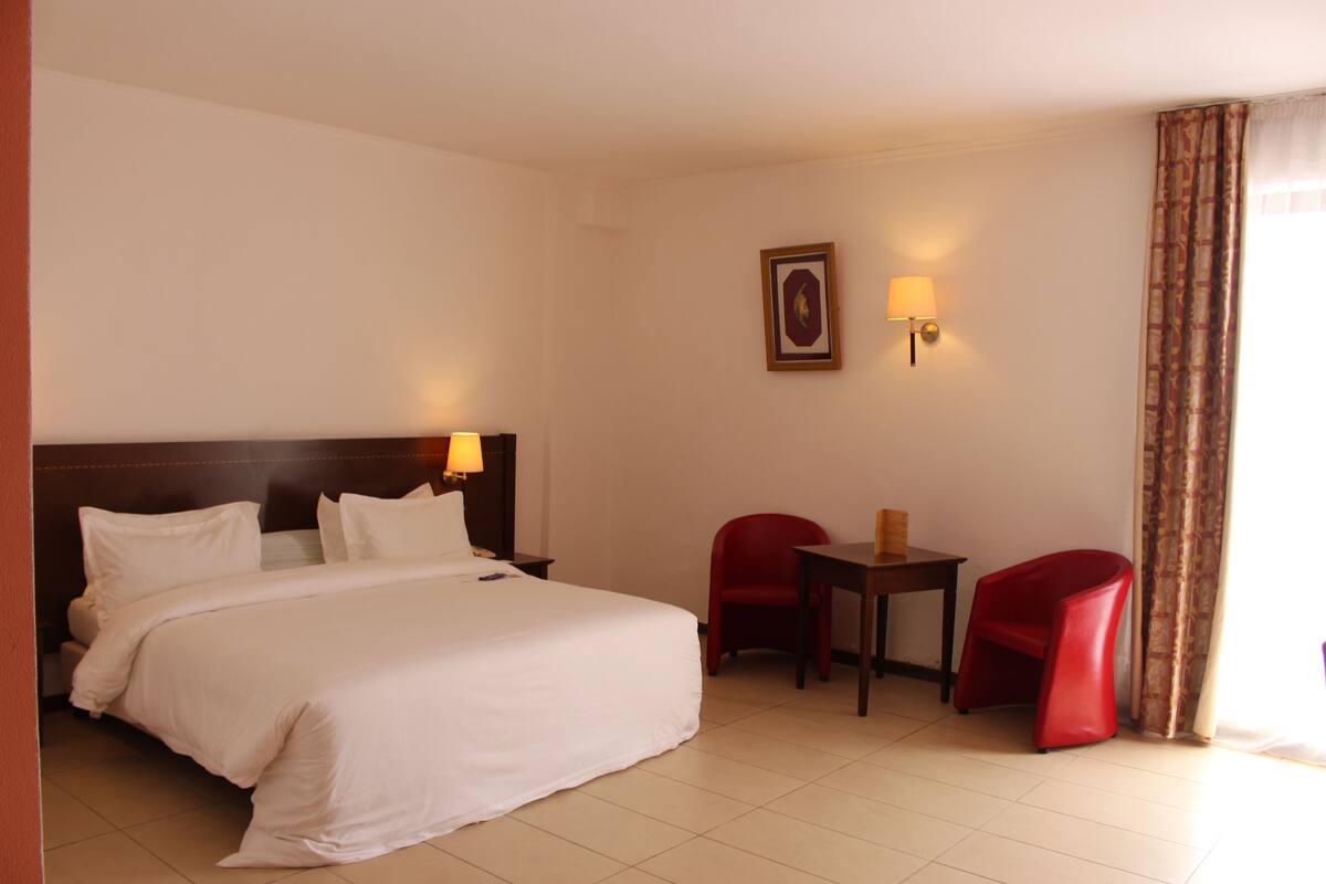 Una habitación de hotel con una cama, dos sillas rojas, una mesita y una lámpara de pared.