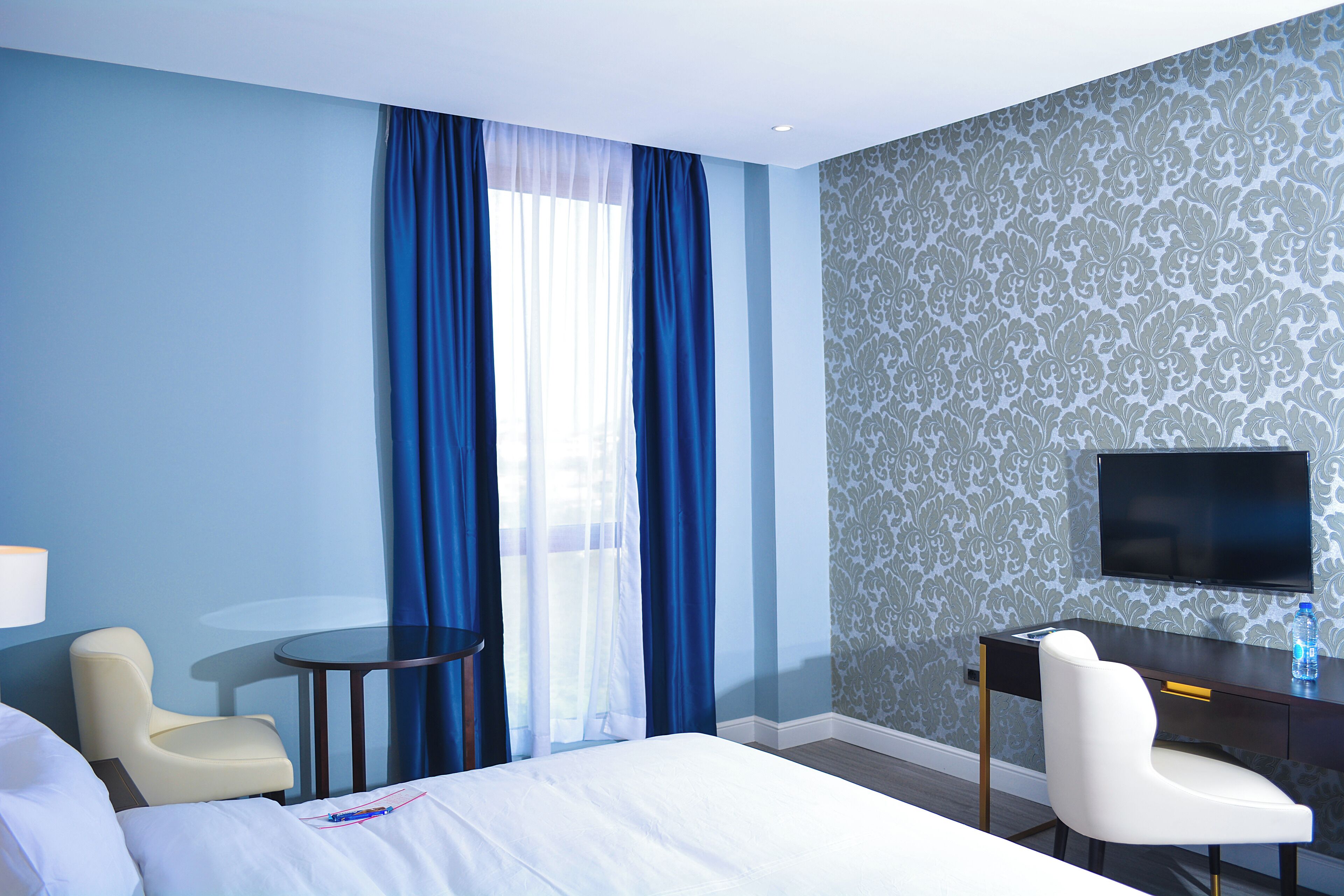 A hotel room with a bed, a desk, a chair, a TV, and a window with blue curtains.