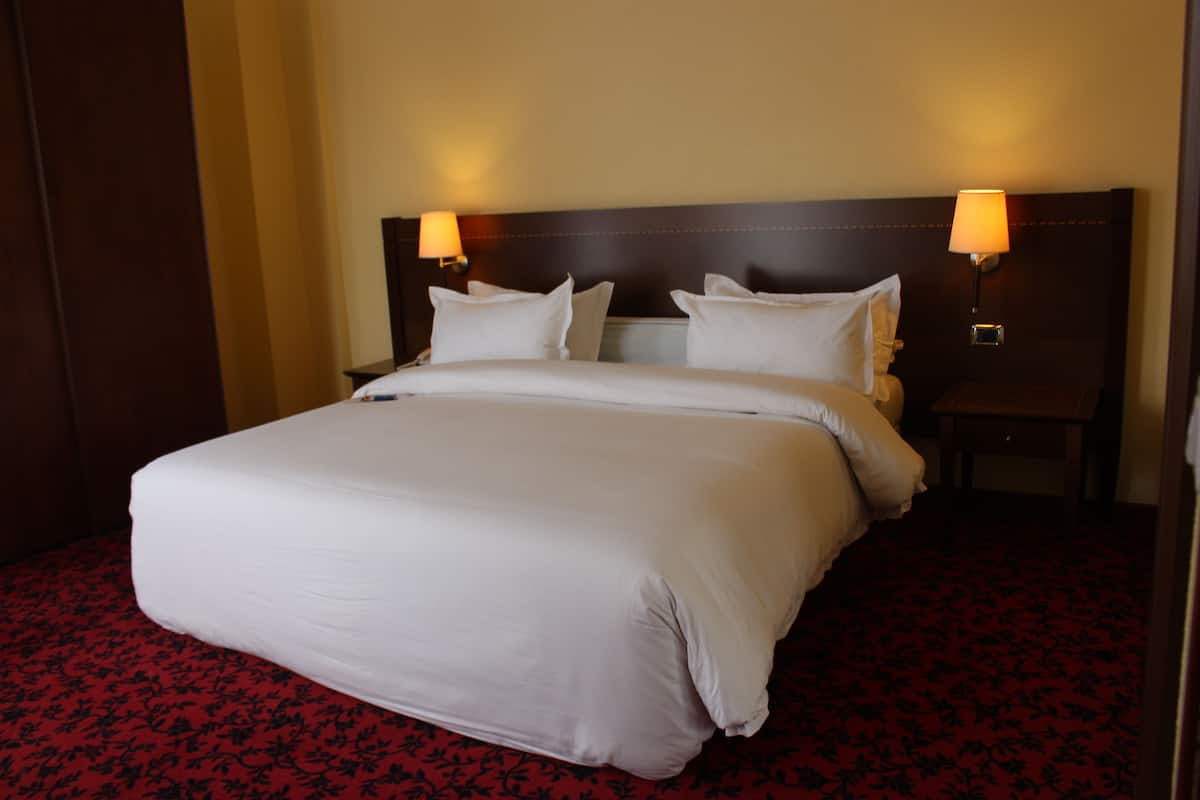 Una cama grande con sábanas blancas, dos mesitas de noche con lámparas y una alfombra roja con estampado floral.