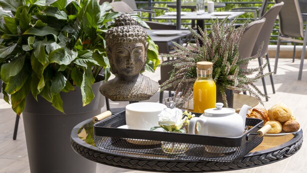 A tray with a Buddha statue, a potted plant, a bottle of orange juice, a teapot, and pastries on an outdoor patio.