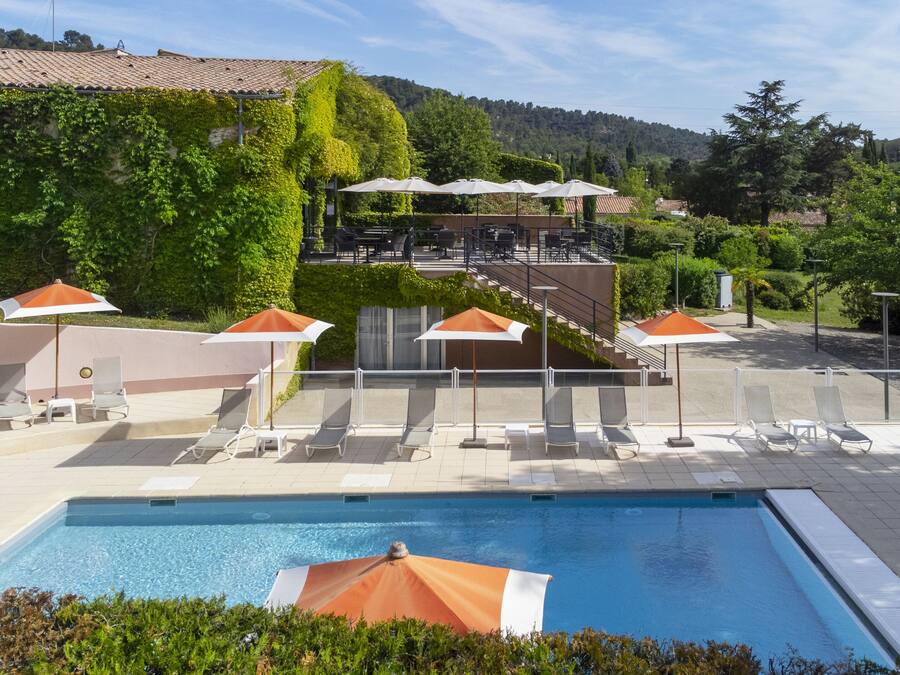 A pool area with lounge chairs and umbrellas, surrounded by greenery and a hillside in the background.
