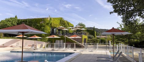 Zona de piscina con sillones de descanso y sombrillas, rodeada de follaje y vistas a una colina.