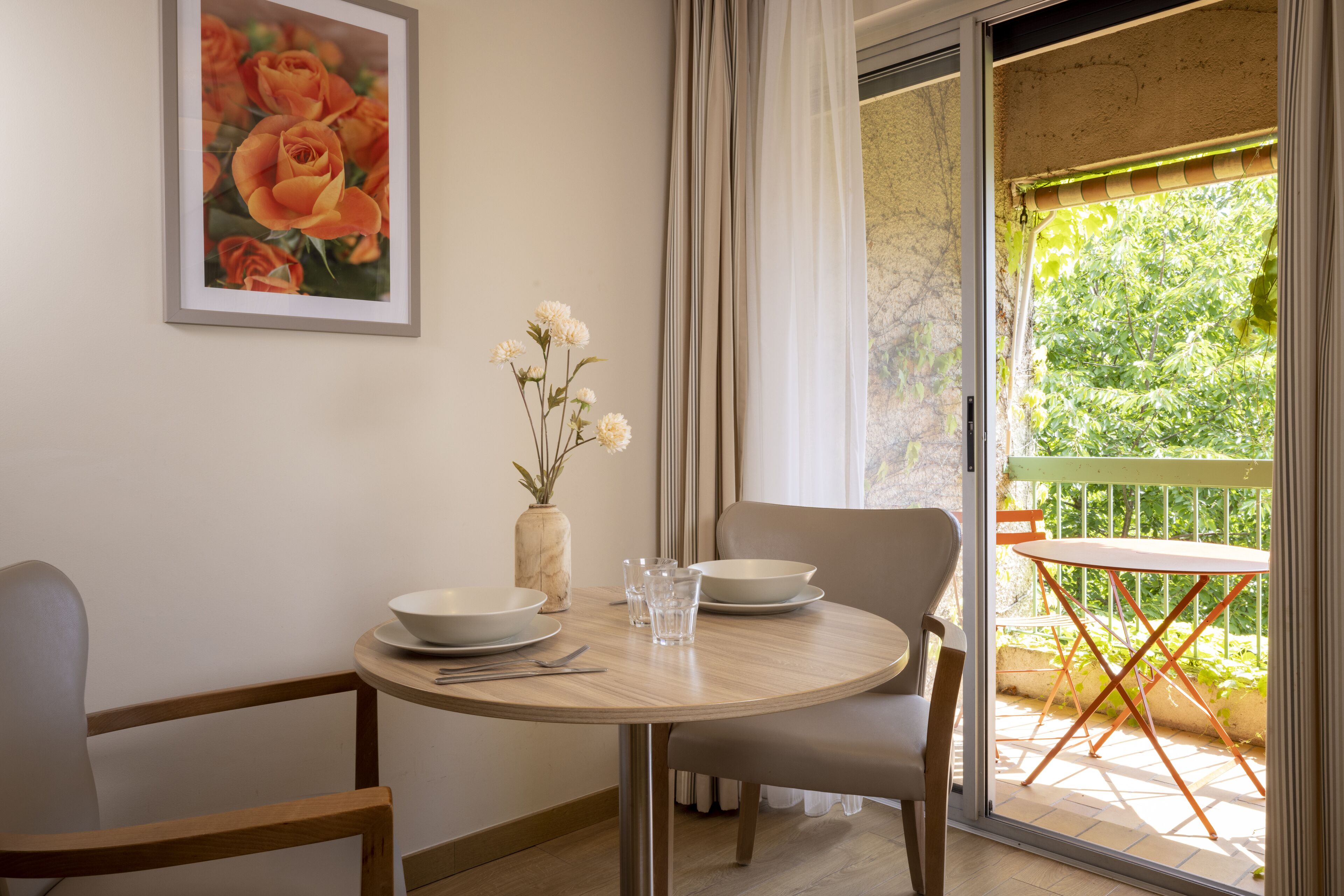 Un comedor con una mesa redonda, sillas y un jarrón con flores. Se observa una ventana con cortinas y vistas al exterior con vegetación.