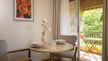 A dining area with a round table, chairs, and a vase with flowers. There is a window with curtains and a view of greenery outside.