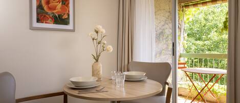 A dining area with a round table, chairs, and a vase with flowers. There is a window with curtains and a view of greenery outside.