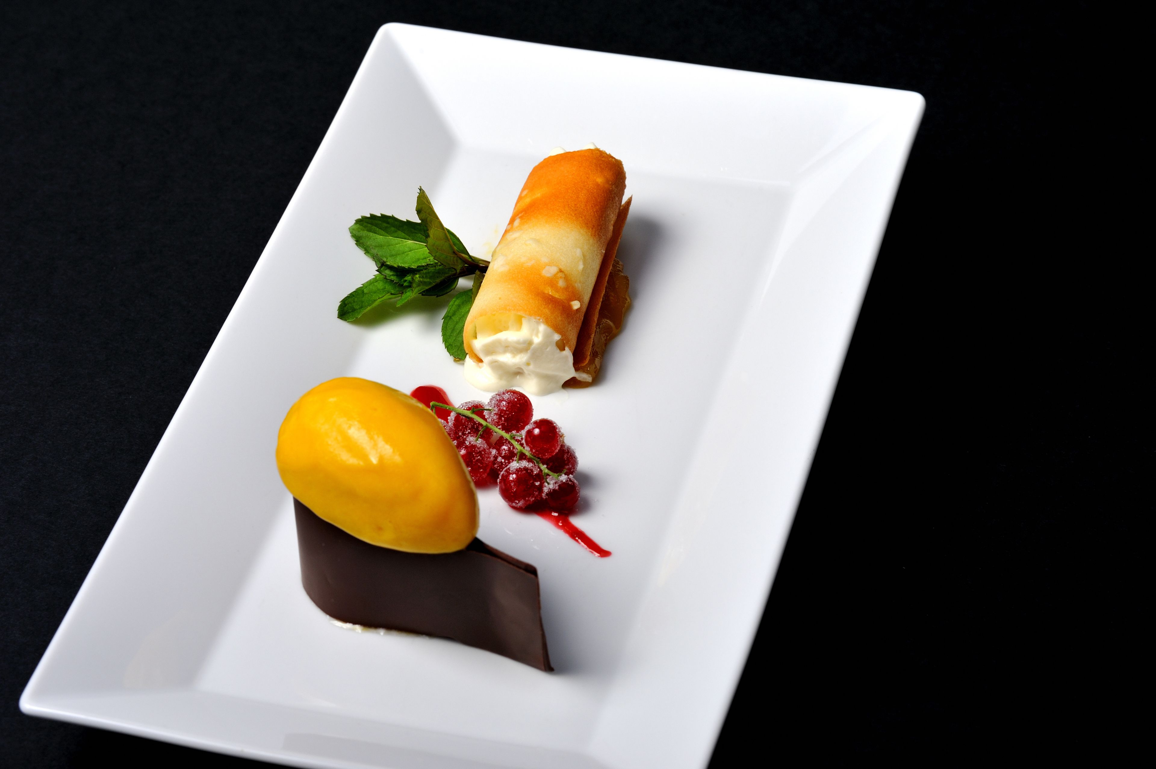 A dessert on a white plate with a chocolate base, a rolled pastry, a yellow fruit, and red berries.