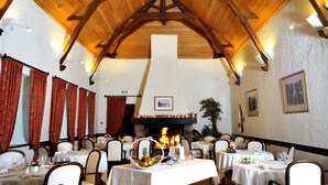 Un restaurant avec des poutres en bois, des murs blancs et une cheminée. Les tables sont dressées avec des nappes blanches et des chaises.