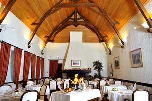 Un restaurante con vigas de madera, paredes blancas y una chimenea. Las mesas están dispuestas con manteles blancos y sillas.