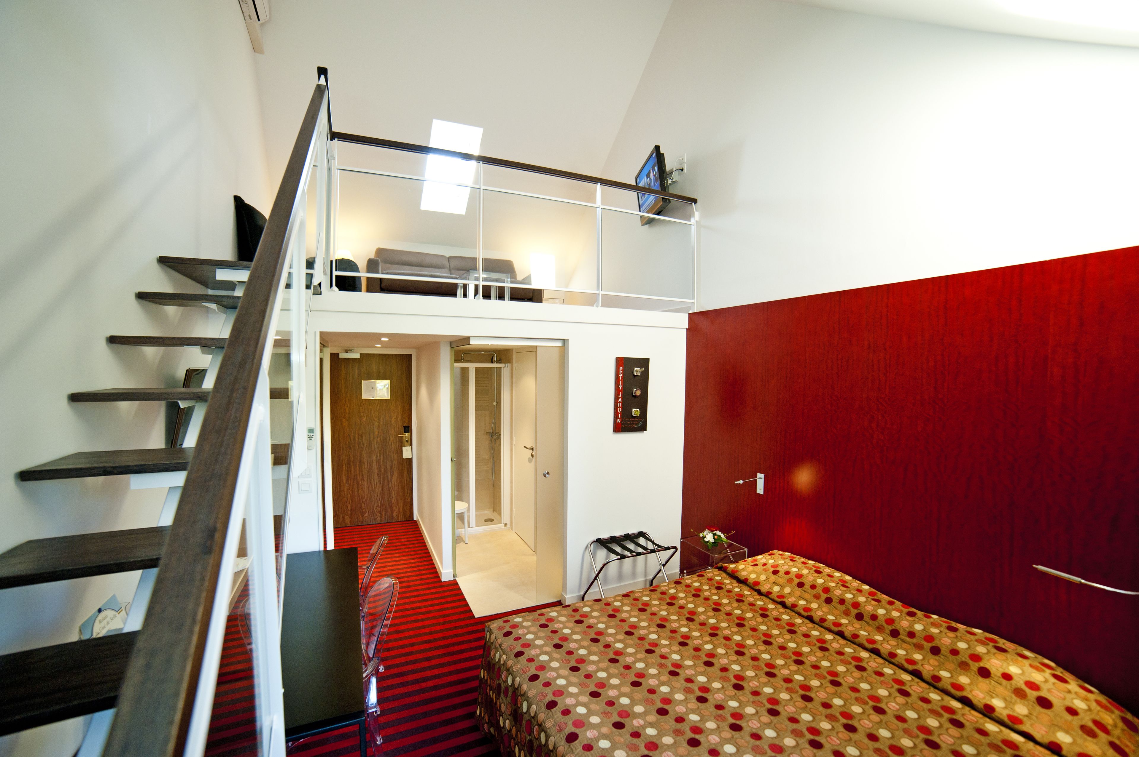 A hotel room with a red wall, a bed with a patterned bedspread, a wooden staircase, and a small table with a chair.