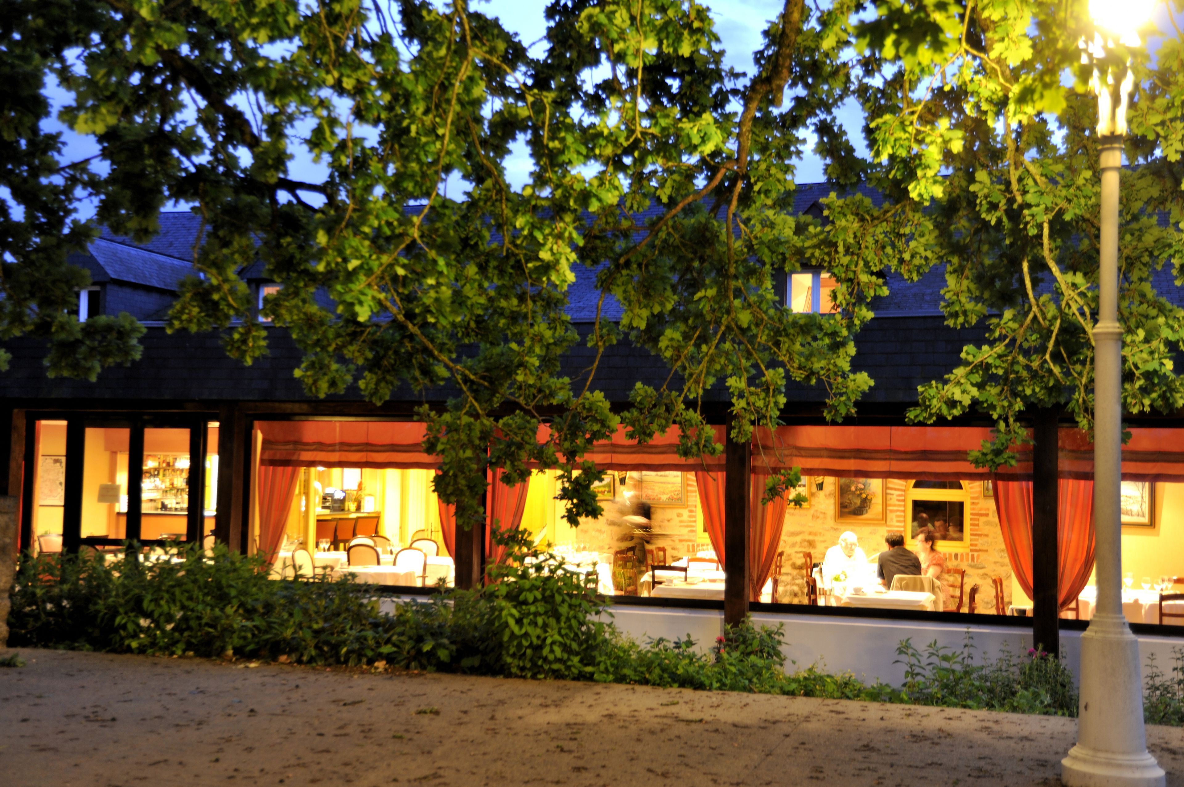A building with a dining area visible through large windows, surrounded by greenery.